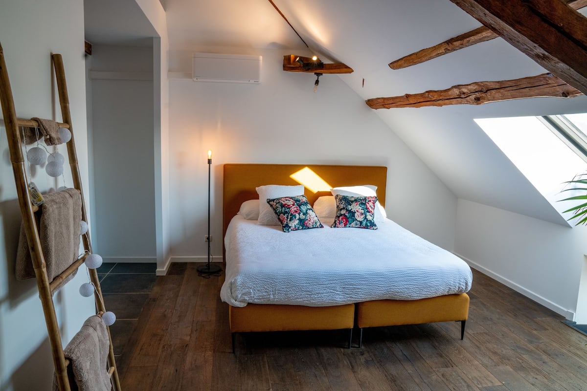 A bedroom with a wooden headboard, a bed with white bedding and floral pillows, a bedside lamp, and a wooden ladder with towels.