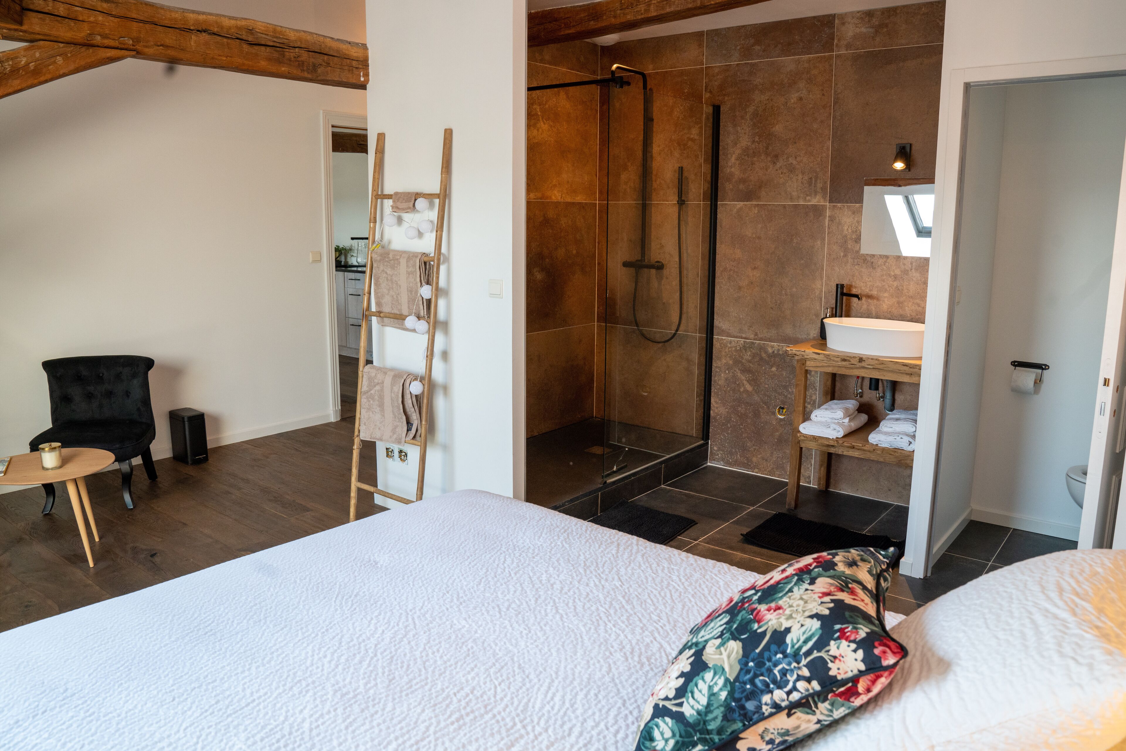 A modern hotel room with a large bed, a shower area, a small table with a chair, and a wooden ladder with towels.