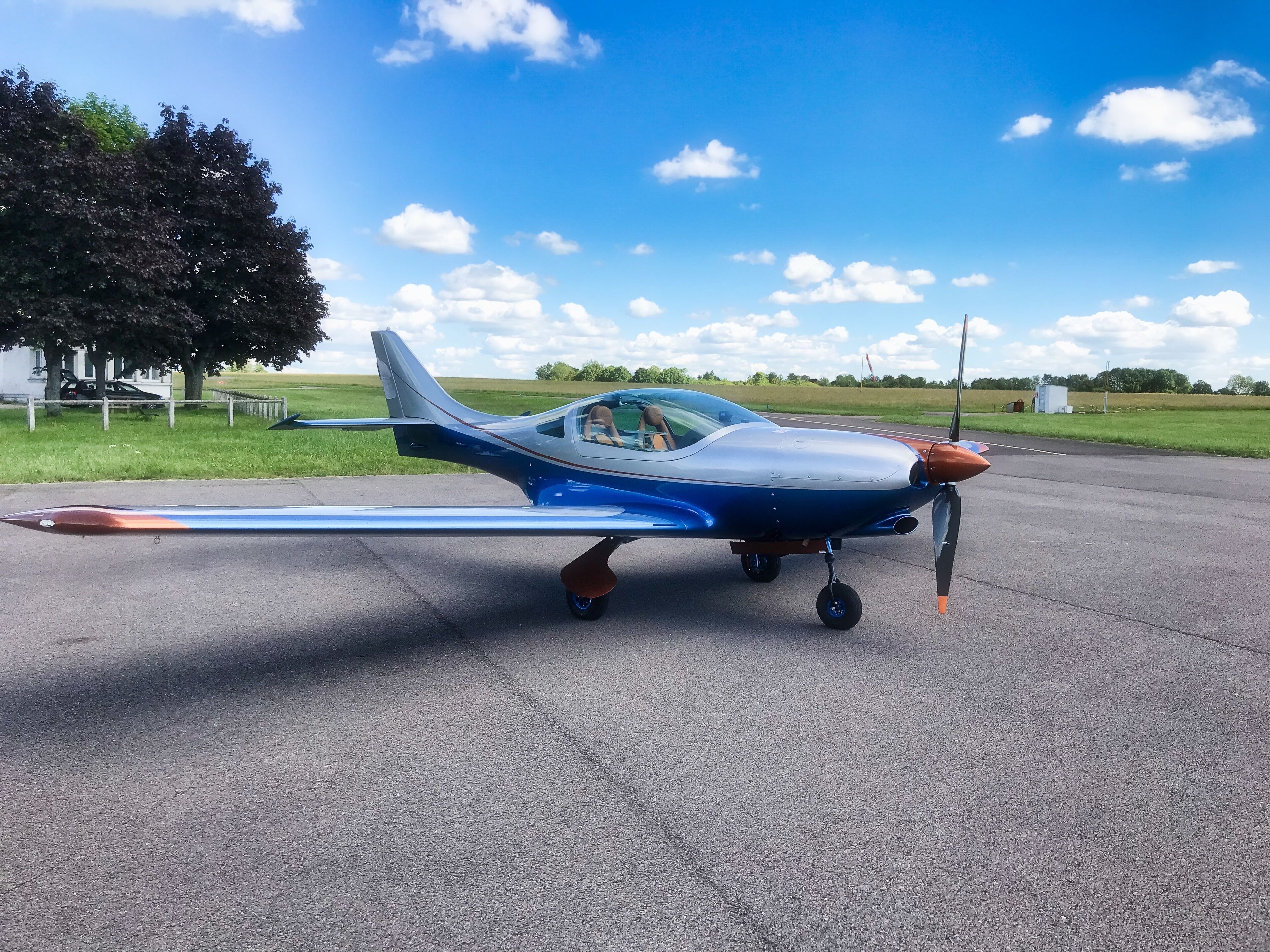 Un petit avion bleu et blanc est garé sur une piste, avec un champ herbeux et des arbres en arrière-plan.