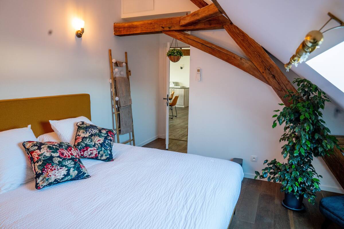 A bedroom with a wooden ceiling, a bed with white bedding, floral pillows, and a potted plant.