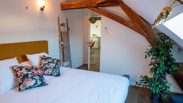 Une chambre avec un plafond en bois, un lit recouvert de draps blancs, des coussins à motifs floraux et une plante en pot.