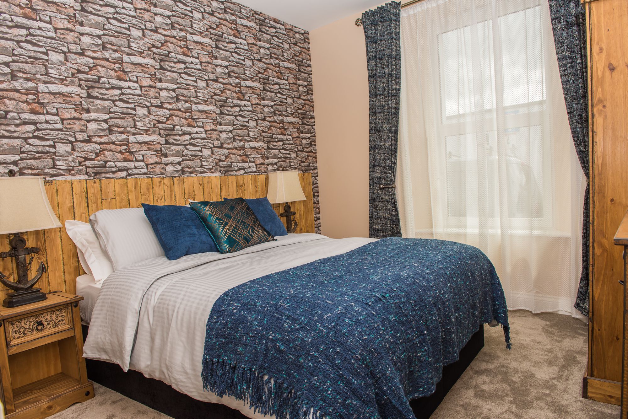 A bedroom with a stone wall, a wooden headboard, a bed with blue bedding, a nightstand with a lamp, and a window with sheer curtains.