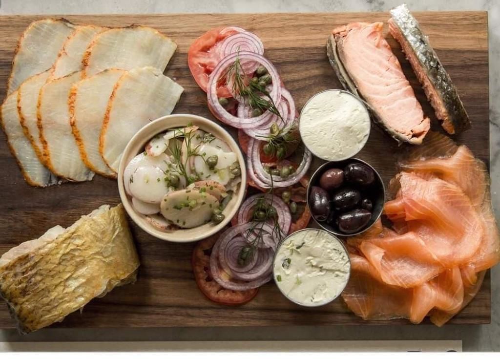 Ein Holzbord mit verschiedenen Lebensmitteln, darunter Fischscheiben, Oliven und eine Salat-Schüssel.