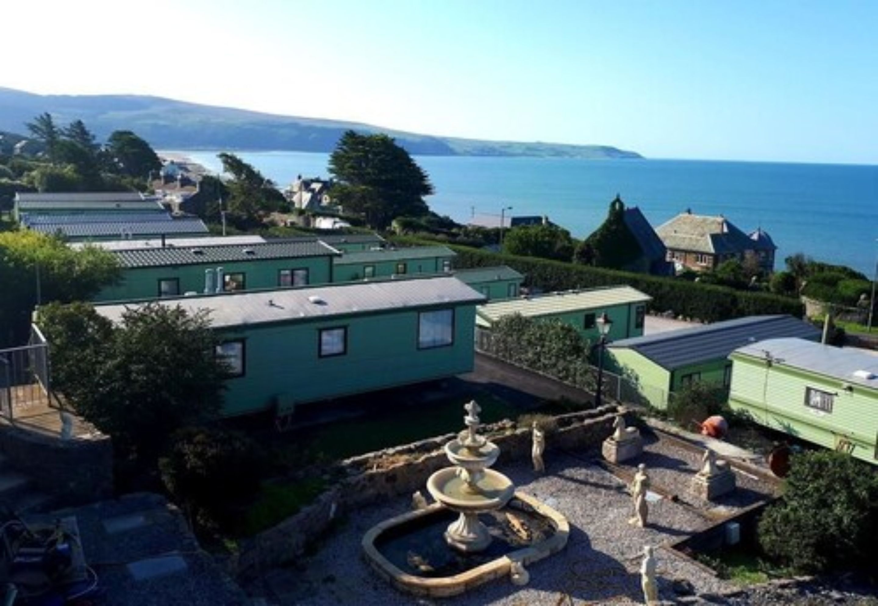 A coastal property with multiple green-roofed buildings, a central fountain, and a view of the sea.