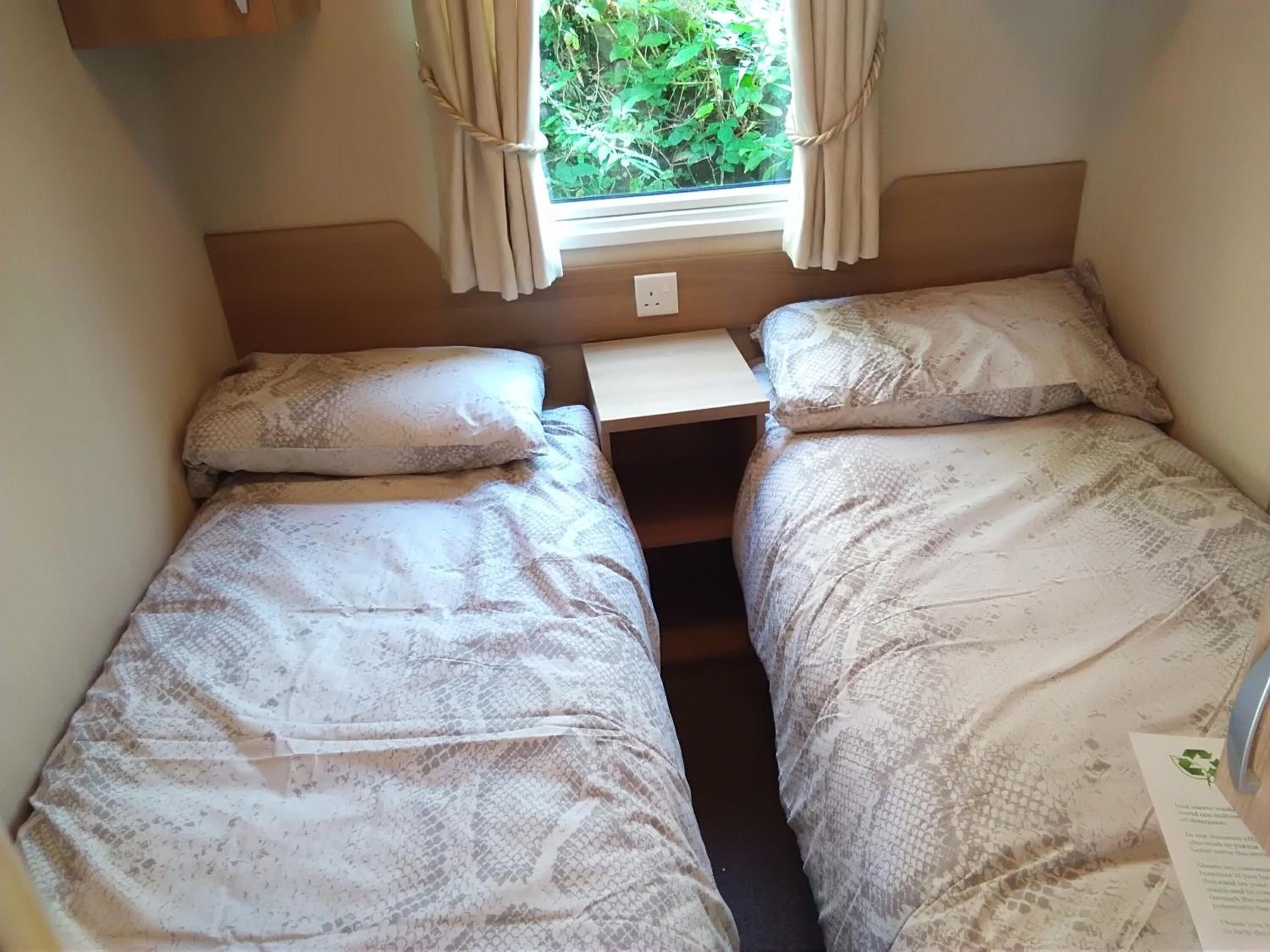 A small, compact bedroom with two single beds, a window with curtains, and a small table.