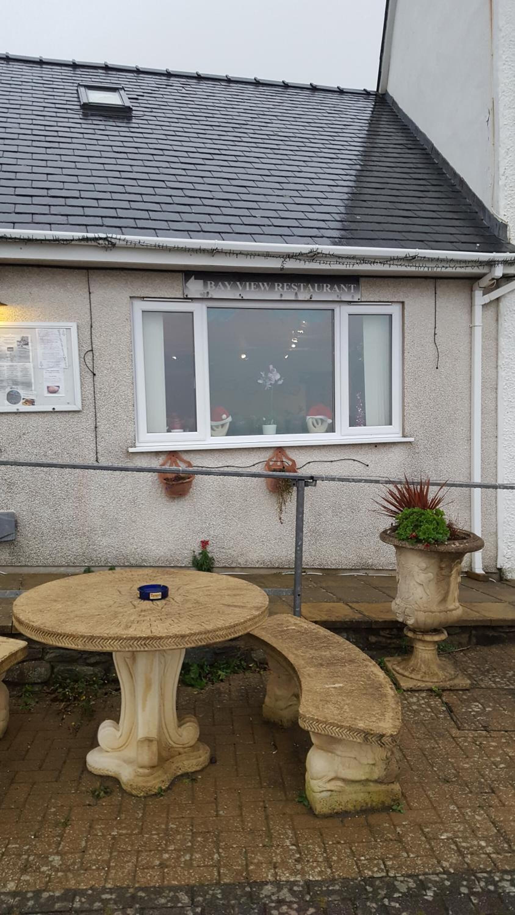 A building with a sign reading "Bay View Restaurant" above a window. There is a round table with chairs in front of the building, and two potted plants are placed on the ground.