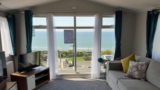 Ein Zimmer mit grauem Sofa, einem Fernseher auf einem Holzboden und Meerblick durch große Glasüren.