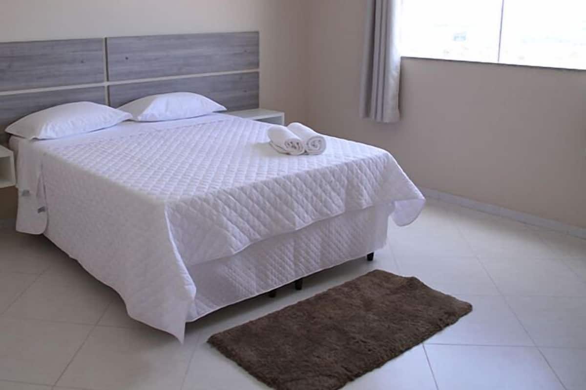 A neatly made bed with white bedding and a brown rug in a bright room with a large window.