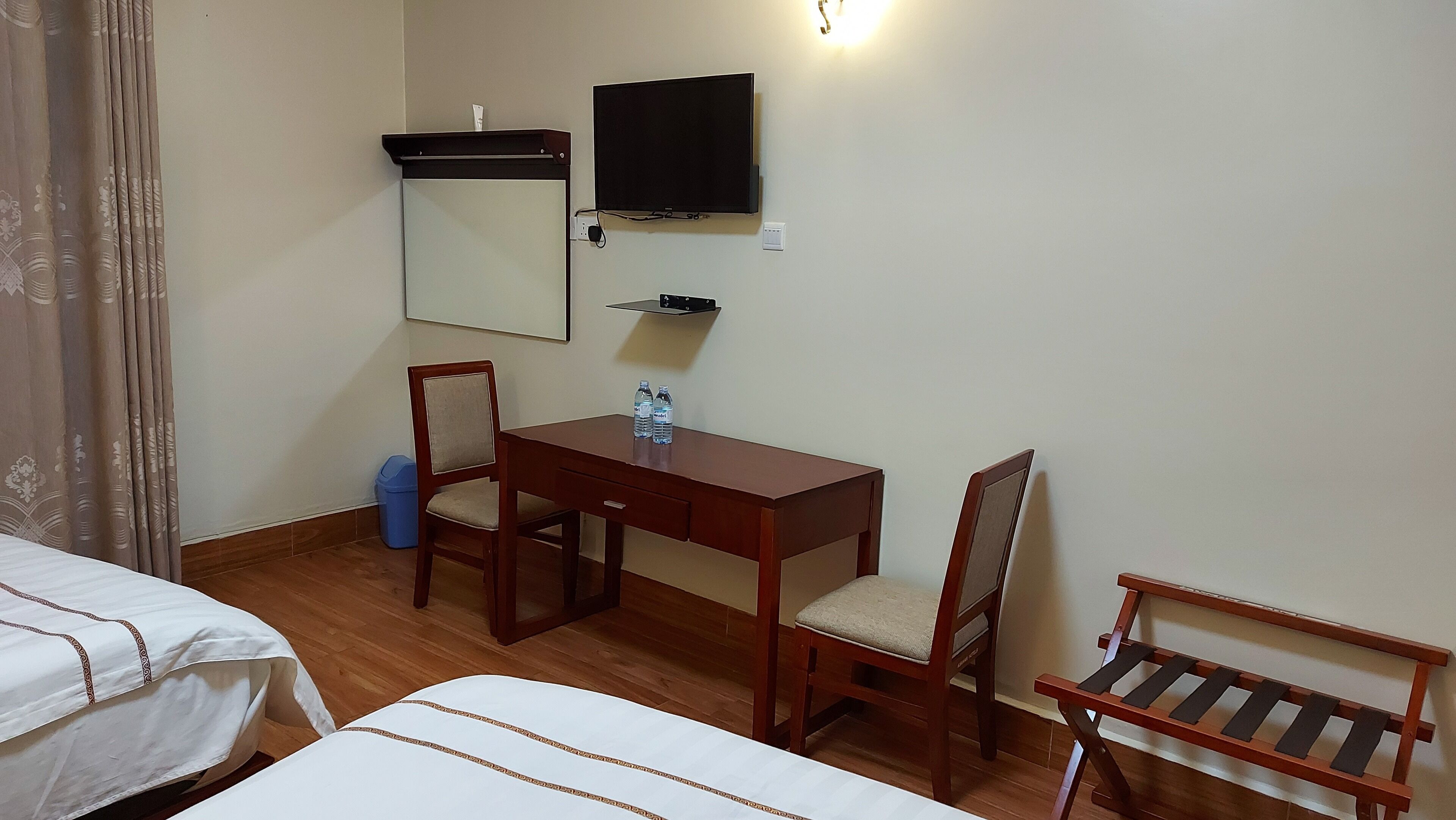 A hotel room with two single beds, a wooden desk with a chair, a television mounted on the wall, and a small shelf below the TV.