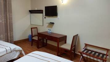 A hotel room with two single beds, a wooden desk with a chair, a television mounted on the wall, and a small shelf below the TV.