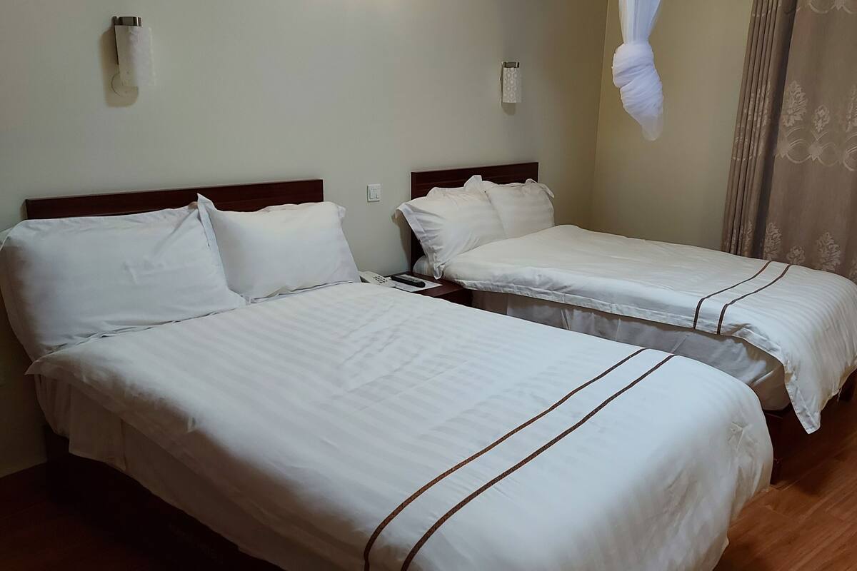 Quarto de hotel com duas camas, cada uma com roupa de cama branca e detalhes em castanho. Possui piso de madeira, cortina na janela e luminária fixada na parede.