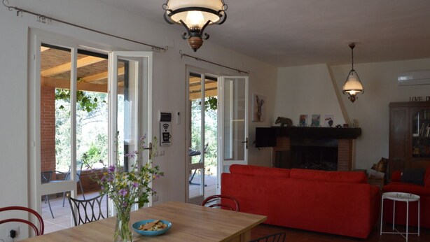 A dining area with a wooden table, chairs, and a vase of flowers. There is a red sofa and a fireplace in the living area. - Cozy vacation home on a farm, Bagno di Gavorrano, Tuscany (Bagno di Gavorrano)