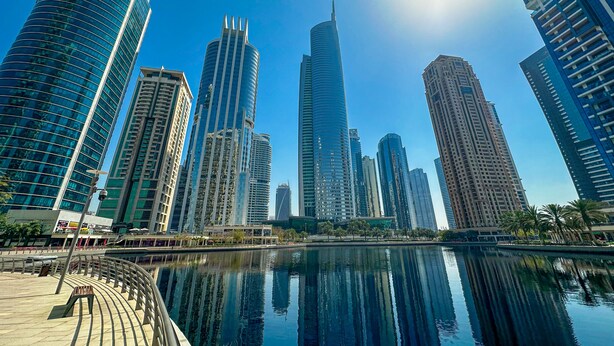 A cityscape with modern skyscrapers and a calm water body reflecting the buildings. - HiGuests - Lake view (Dubai)