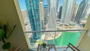 A balcony with a view of a cityscape, including tall buildings and a body of water. There is a wooden table with two chairs and a potted plant. - HiGuests - Lake view (Dubai)