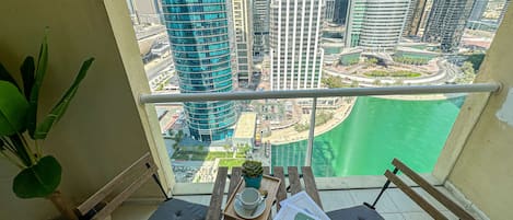 A balcony with a view of a cityscape, including tall buildings and a body of water. There is a wooden table with two chairs and a potted plant.