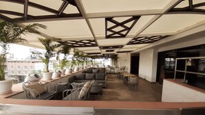 A modern lounge area with a long sofa, chairs, and tables, surrounded by potted plants and a view of buildings. - Hotel 18 Keys (Bhopal)