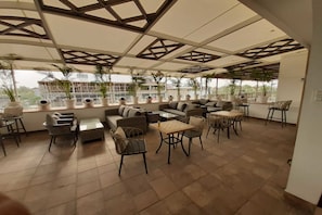 A spacious outdoor seating area with multiple sofas, chairs, and tables, surrounded by large windows and potted plants.