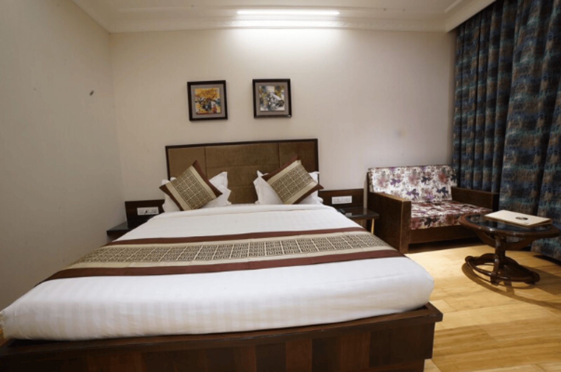 A neatly arranged hotel room with a large bed, a wooden headboard, a sofa, and two framed artworks on the wall.