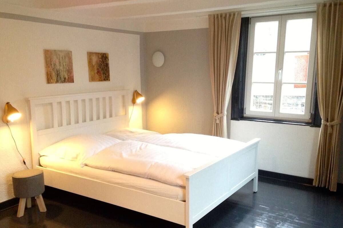 A neatly made bed with white bedding, two wall-mounted lamps, and a window with curtains.