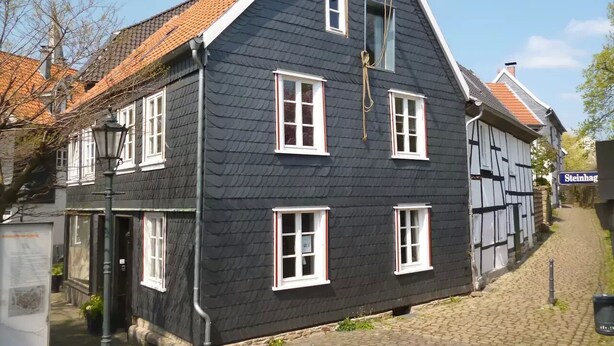 Ein traditionelles, schwarzes Gebäude mit weißen Fensterrahmen und einem geneigten Dach, an einer gepflasterten Straße gelegen.