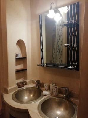 A bathroom with two sinks, a mirror with a wavy frame, and a shelf with toiletries. - Villa River PALM-PALMERAIE-VLC-263 (Marrakech)