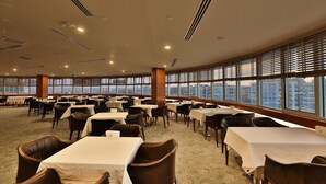 Un amplio comedor con varias mesas y sillas, ventanas grandes con persianas y vistas a la ciudad.
