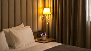 Una habitación de hotel con cama, mesita de noche, lámpara y un amplio ventanal con cortinas.