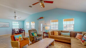 A spacious living room with a high ceiling fan, a dining area with a table and chairs, and a large window with blinds. - Private Pool, Pet Friendly, Golf Cart Access, Close to Beach (Port Aransas)