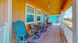 A covered porch with wooden chairs and tables, a teal-colored building in the background, and a clear view of the street. - Private Pool, Pet Friendly, Golf Cart Access, Close to Beach (Port Aransas)
