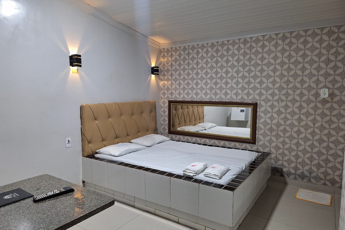A neatly made bed with a headboard, a bedside table with a remote control, and a mirror above the bed.