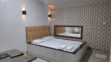 Una cama bien hecha con cabecera, una mesita de noche con control remoto y un espejo sobre la cama.