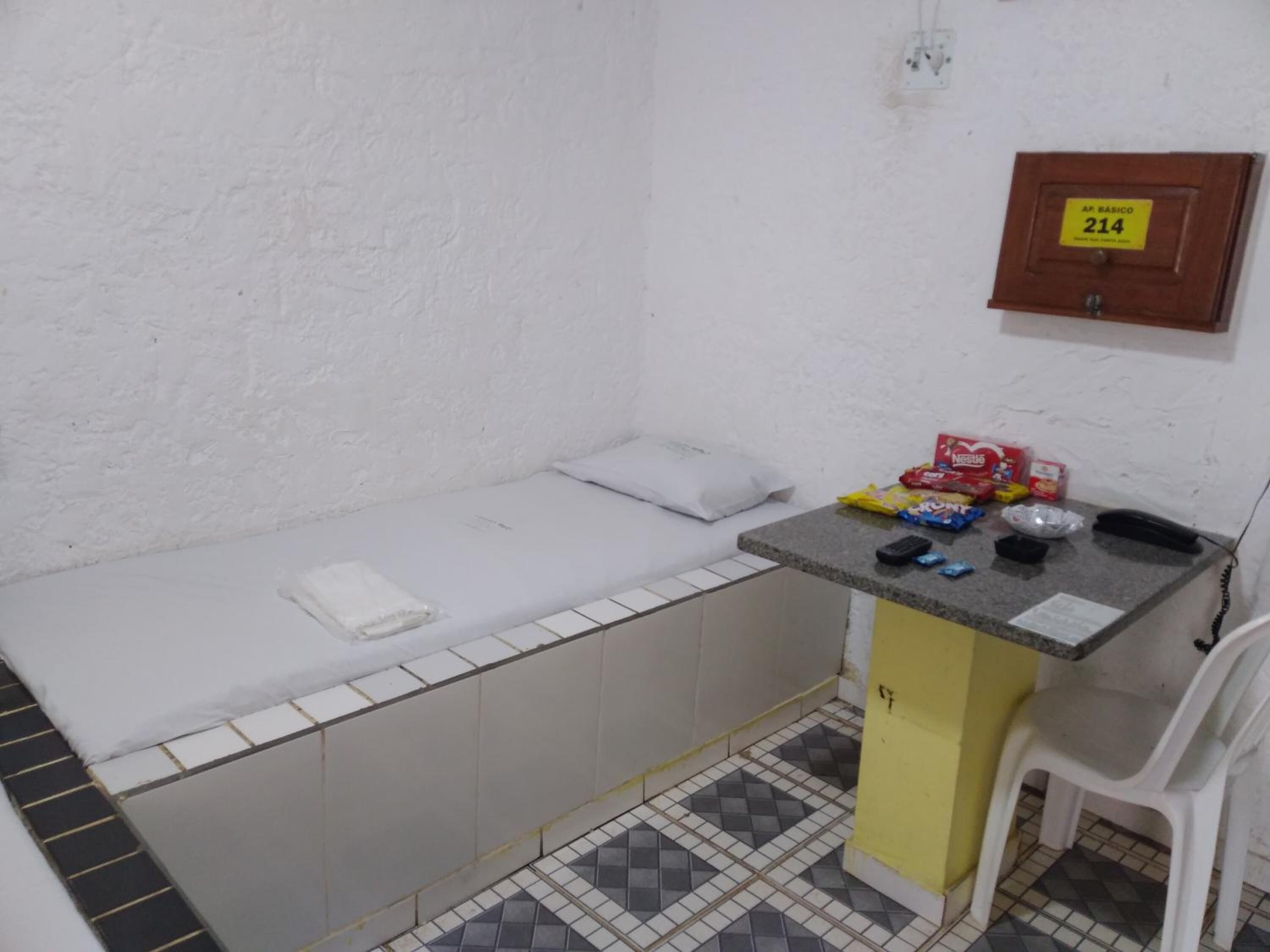 A small, basic room with a single bed, a small table with toiletries, a chair, and a wall-mounted phone.