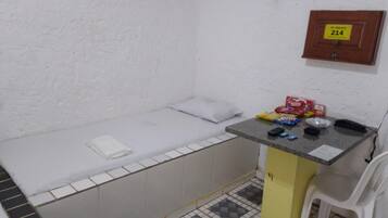 A small, basic room with a single bed, a small table with toiletries, a chair, and a wall-mounted phone.