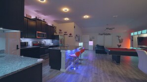Una cocina moderna con armarios oscuros, una isla central con taburetes de bar, mesa de billar y televisor de pantalla plana.