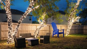 A backyard with a wooden fence, a blue Adirondack chair, wicker furniture, and string lights on trees.