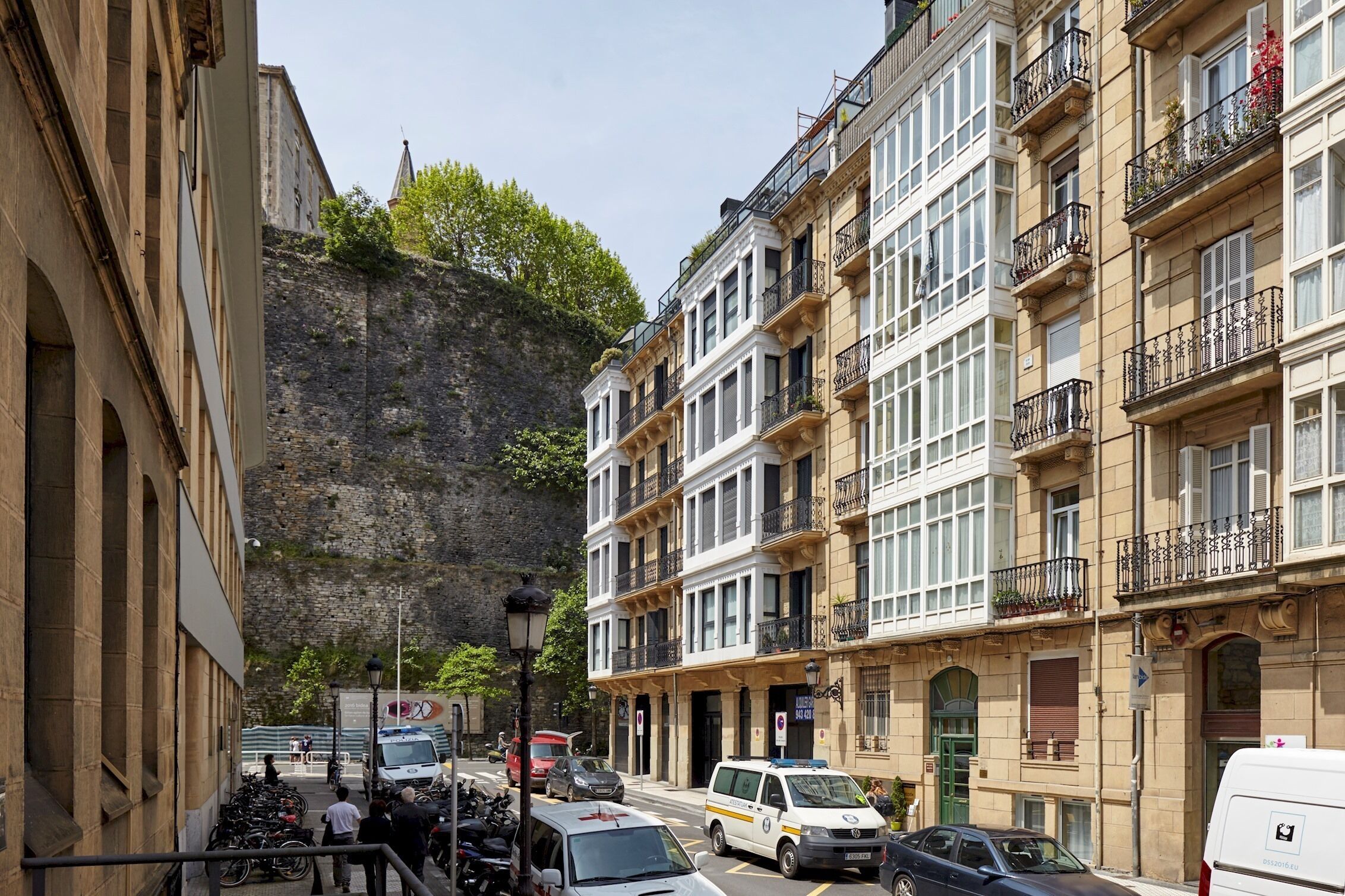A street view with a building featuring multiple floors, balconies, and a stone wall in the background.