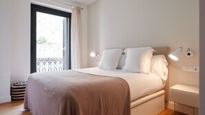Un lit bien fait, recouvert d’une couverture marron, une table de chevet avec une lampe et une fenêtre donnant sur un balcon.