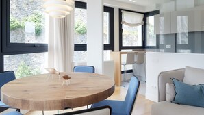 Un salon moderne avec une table ronde en bois, des chaises bleues, un canapé avec des coussins bleus et de grandes fenêtres donnant sur un paysage verdoyant.