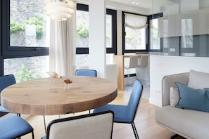 A modern living room with a round wooden table, blue chairs, a sofa with blue cushions, and large windows with a view of greenery.