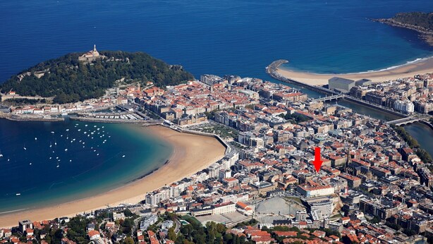 Une vue aérienne d’une ville côtière, avec une imposante forteresse perchée sur une colline, une plage et une zone urbaine dense, une flèche rouge indiquant un bâtiment en particulier.