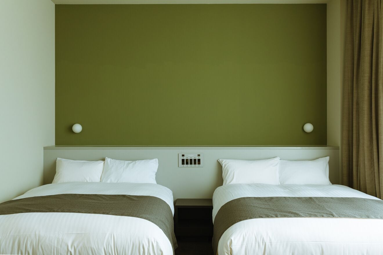 A hotel room with two single beds, a headboard with a light switch, and a wall-mounted air conditioning unit.