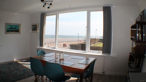 A dining area with a table and chairs, a view of the beach, and a bookshelf. - Eastwatch Guesthouse (Berwick-upon-Tweed)