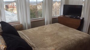 Een slaapkamer met een bed, een televisie op een houten dressoir en een raam met gordijnen.
