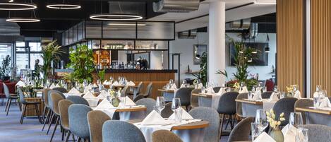 A modern restaurant with round tables, grey chairs, and a bar area in the background.