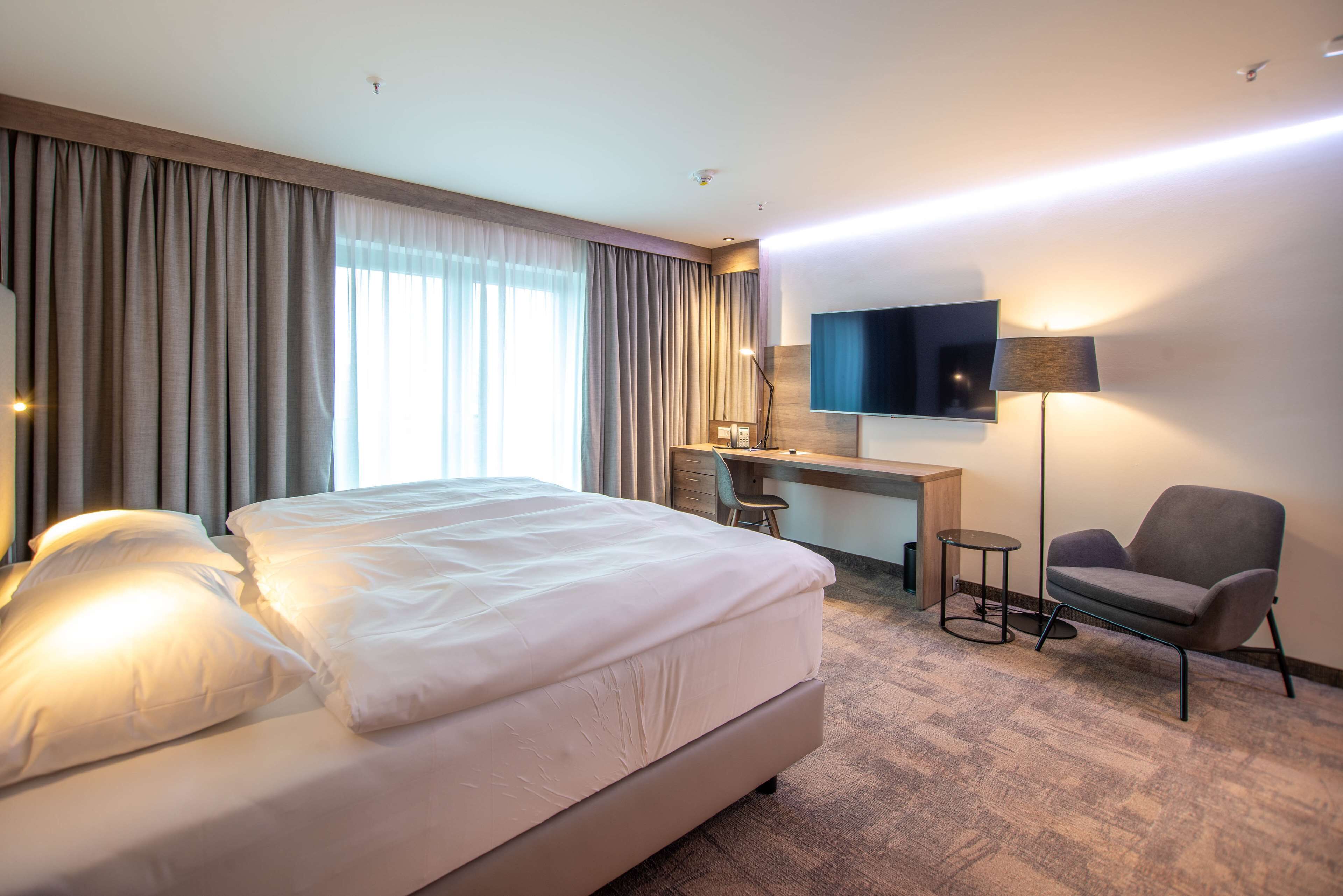 Habitación de hotel con una cama grande, un escritorio con silla, un televisor en la pared y una lámpara.