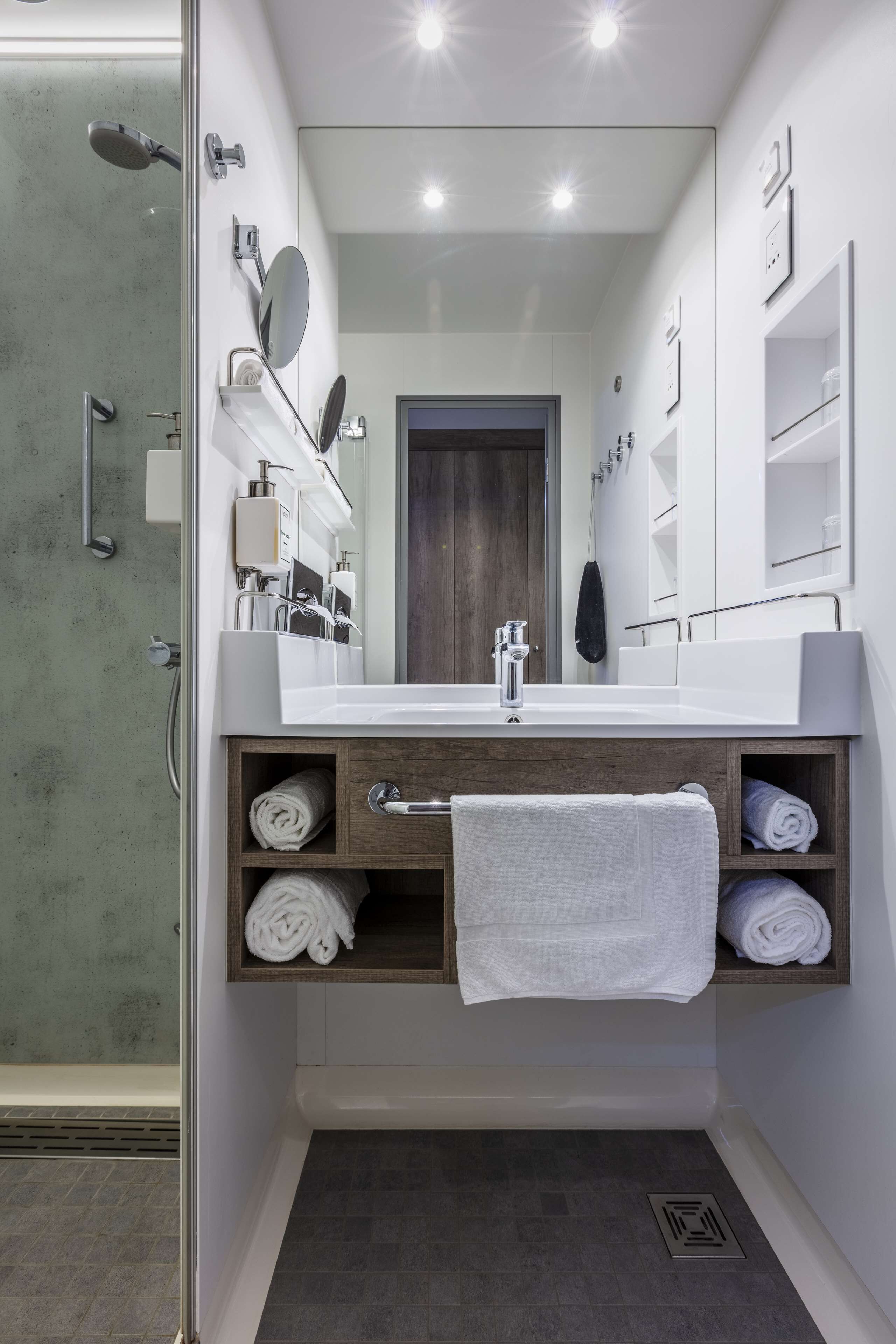 Une salle de bain moderne avec une douche à parois de verre, un lavabo blanc avec un miroir et des serviettes soigneusement pliées, rangées sur des étagères.