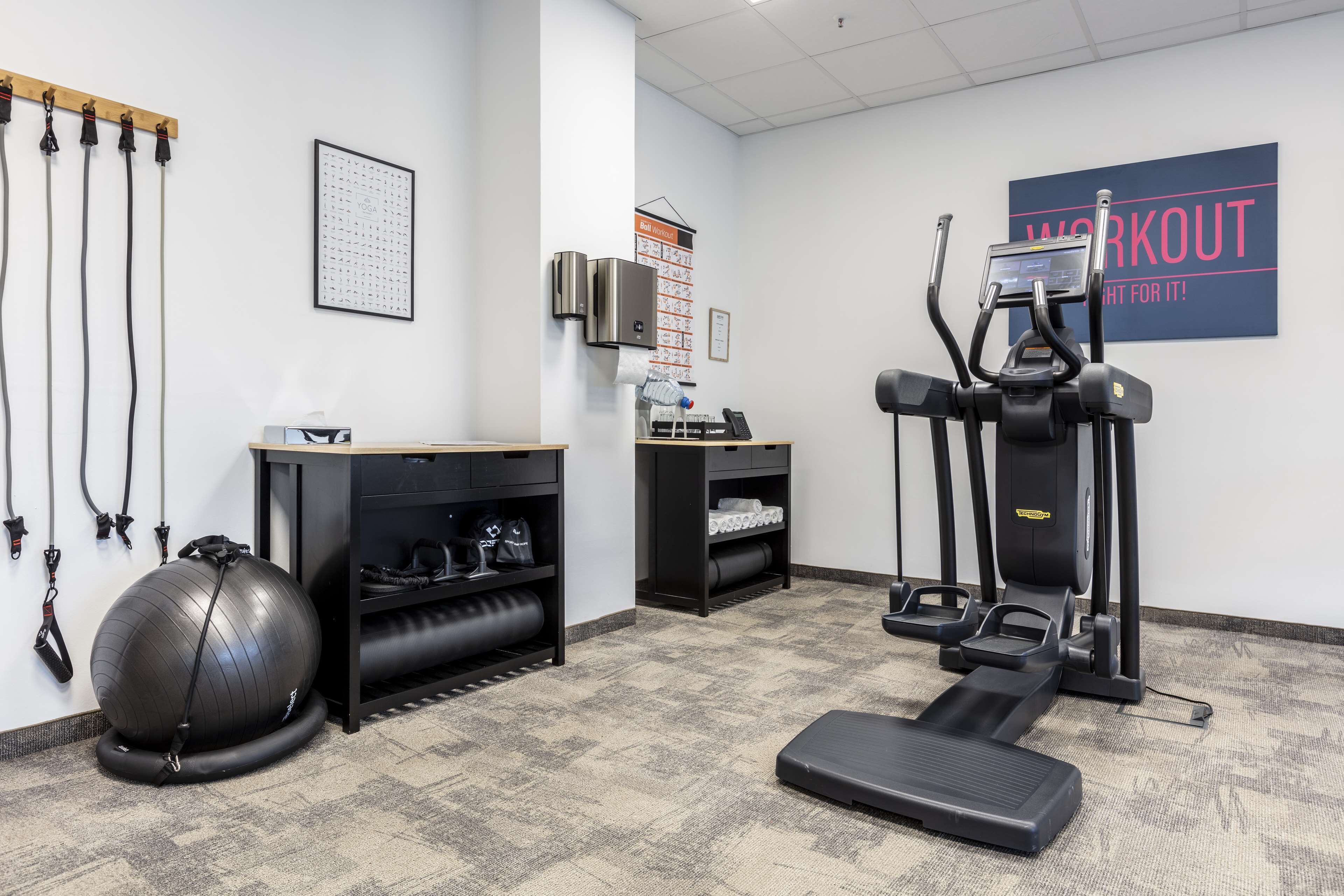 A fitness room with exercise equipment, a wall-mounted air purifier, and a bulletin board with workout schedules.