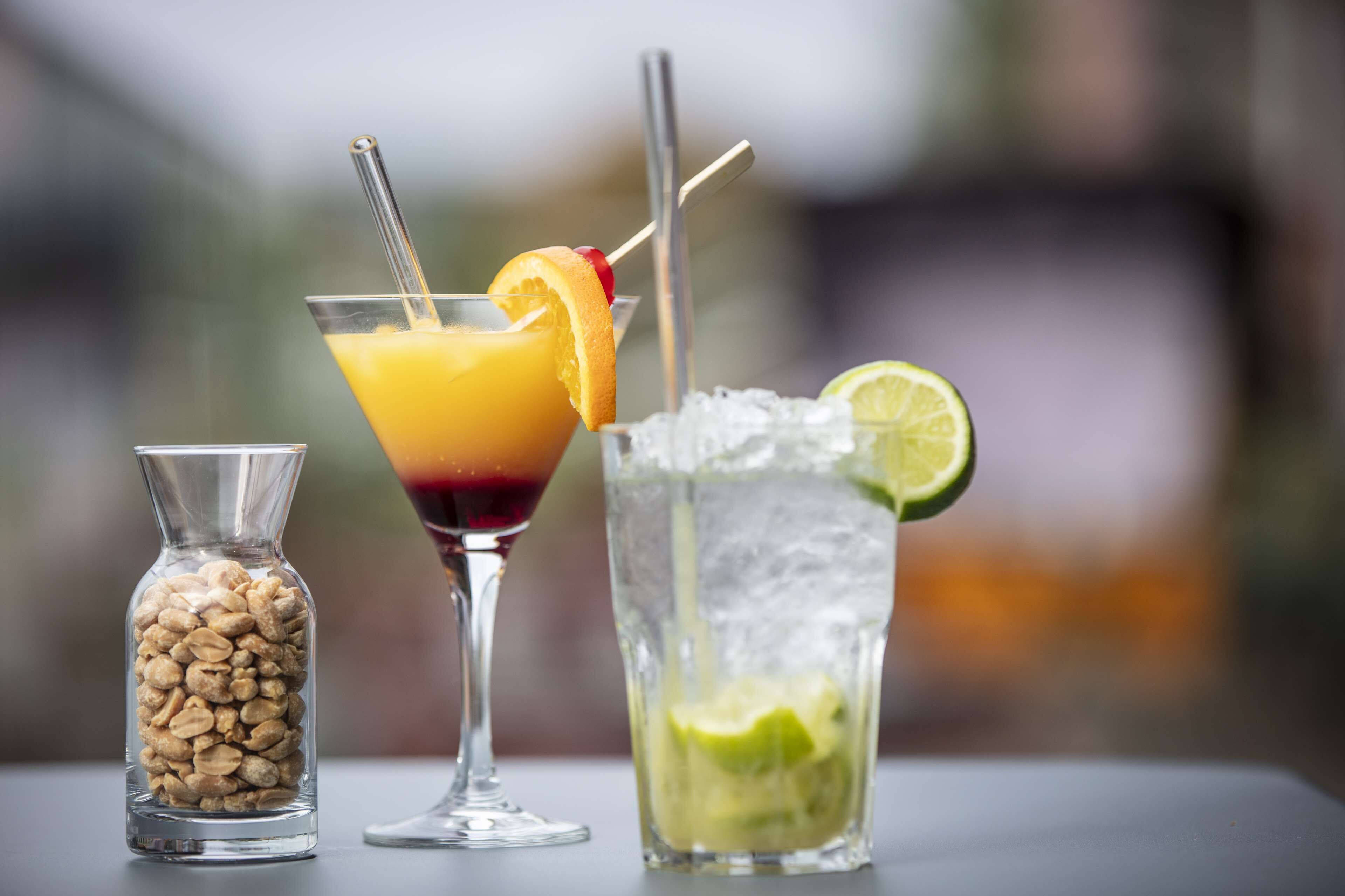 Three drinks on a table: a cocktail with an orange slice and straw, a drink with a lime slice and straw, and a glass with ice and lime.
