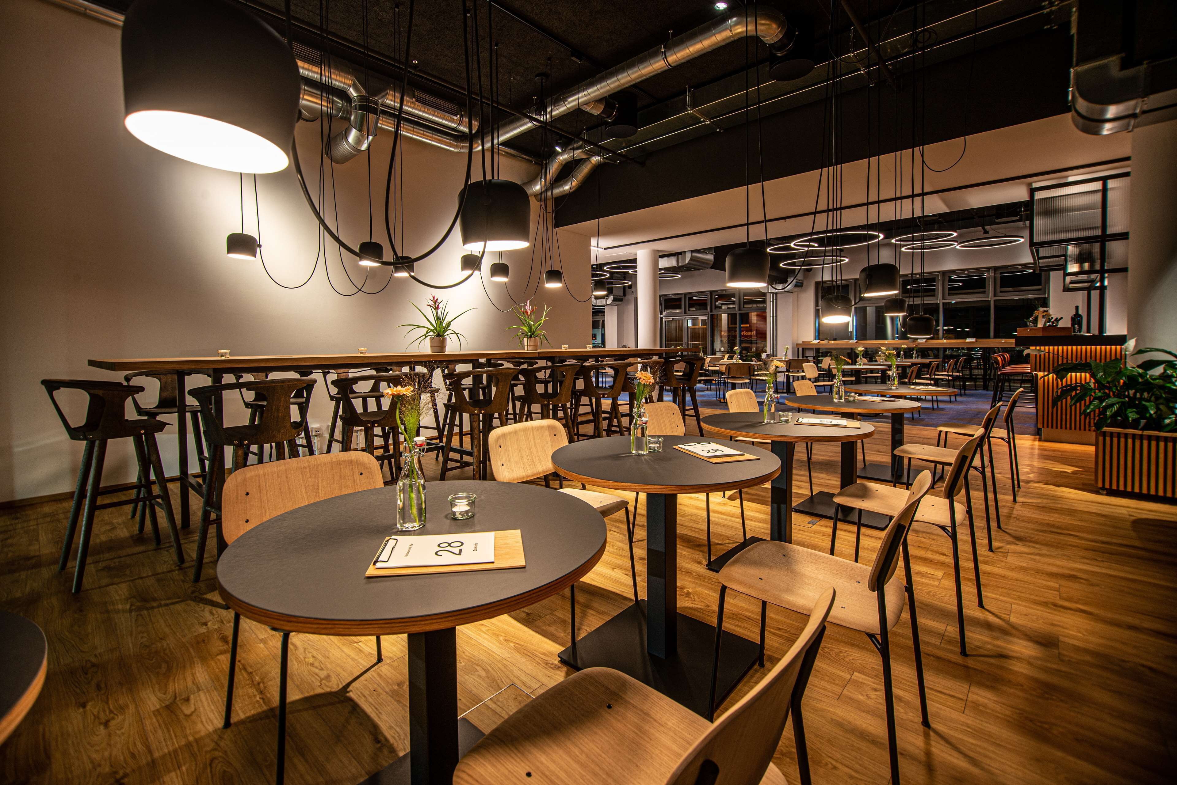 Un restaurant moderne avec des tables rondes et des chaises en bois, agrémenté de luminaires suspendus et de plantes en pot.