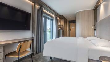 A modern hotel room with a large bed, a desk with a chair, a flat-screen TV mounted on the wall, and a view of the outdoors through a large window.